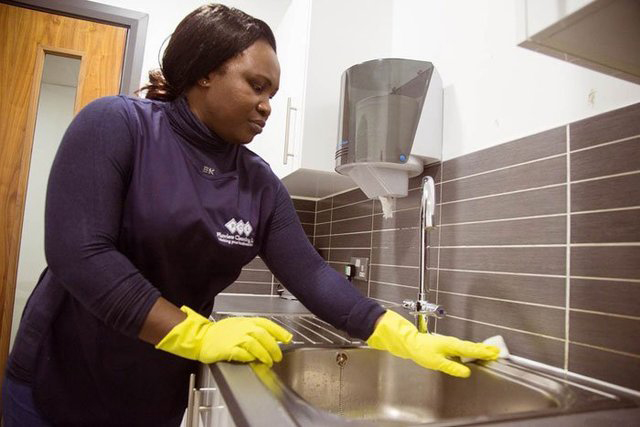 Woman with yellow gloves and Flawless Cleaning Ltd uniform washing around a steel sink